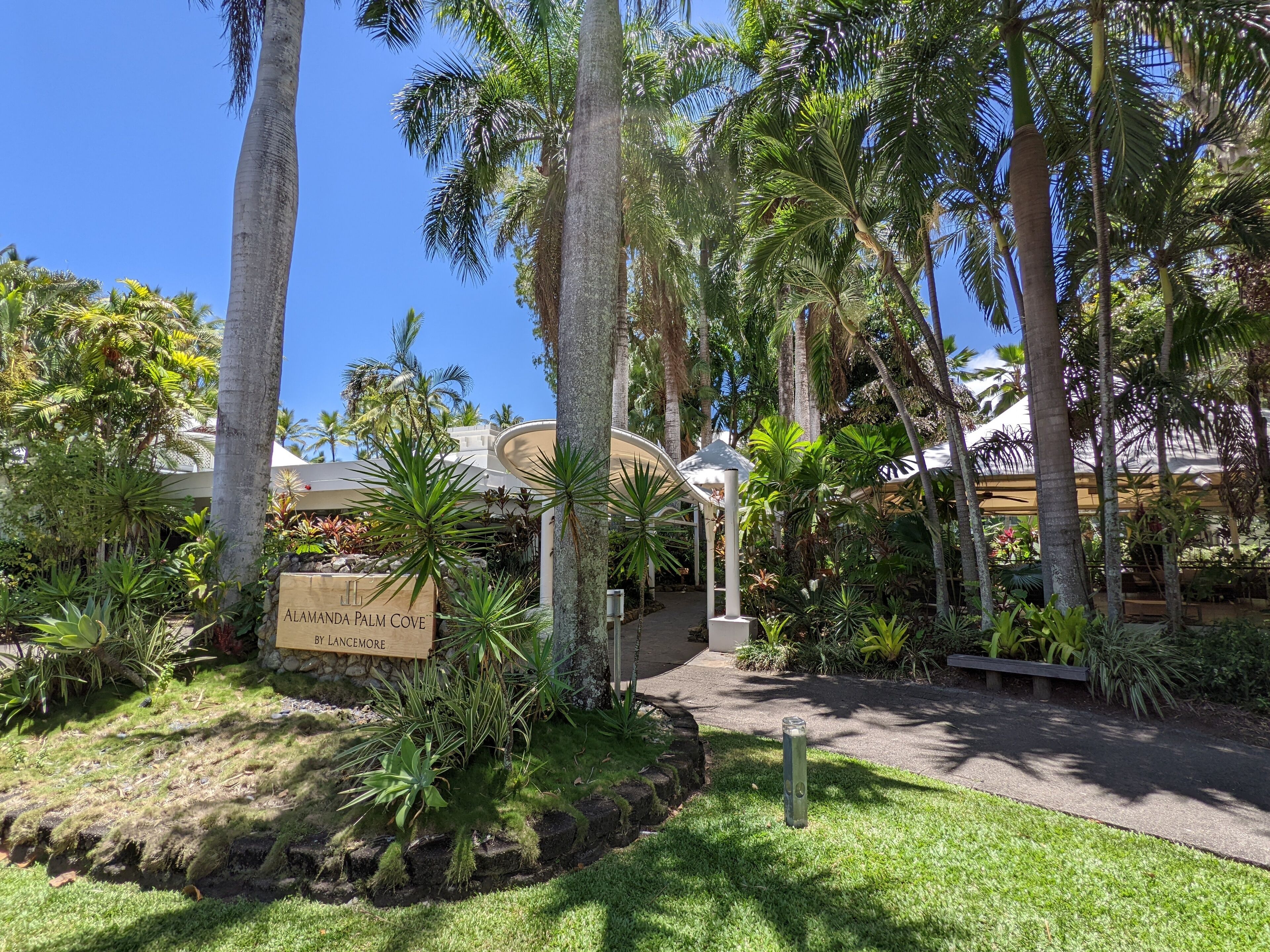 A tropical garden with palm trees, a sign reading "Alameda Palm Cove by Lancimore," and a pathway leading to a building.
