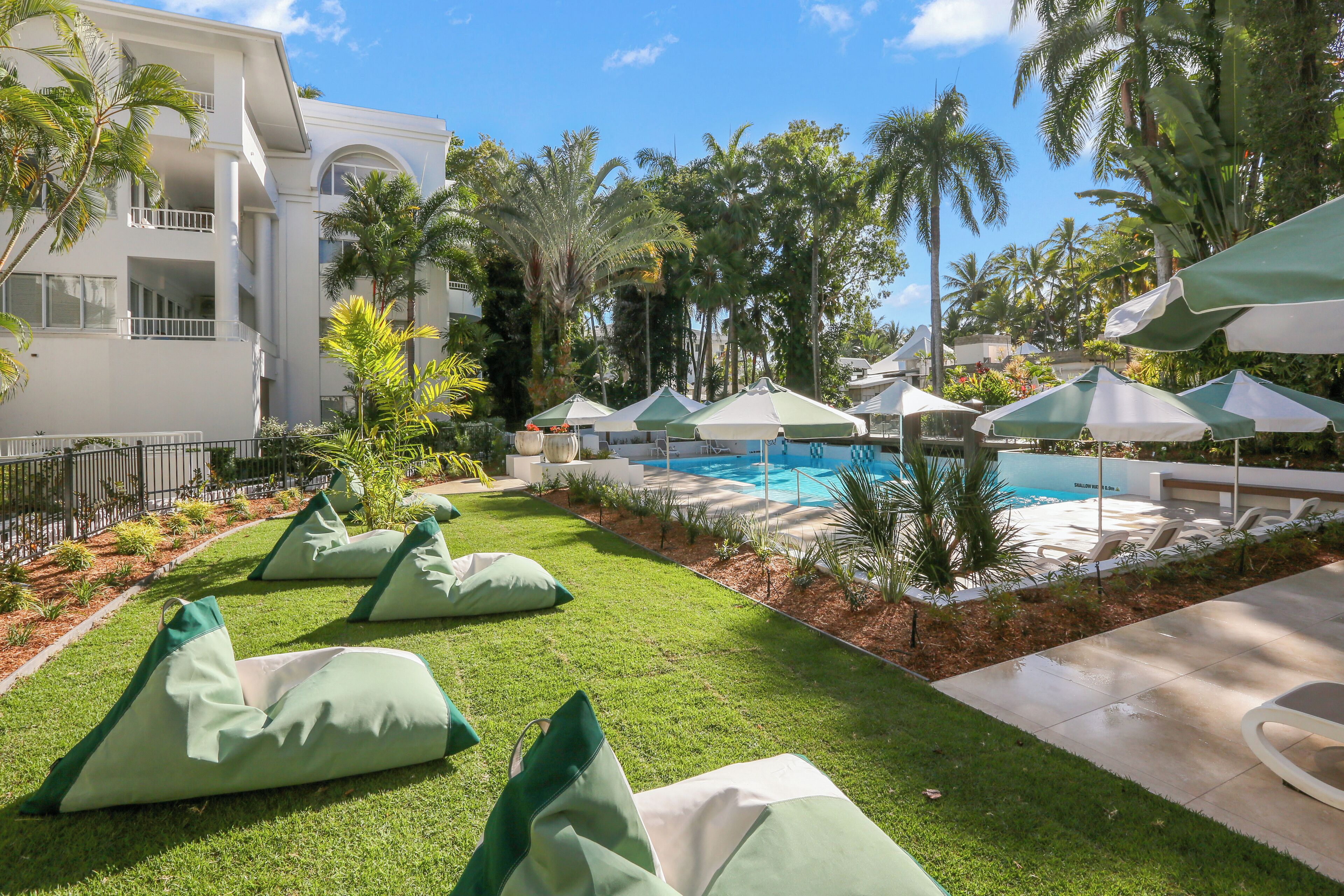 A pool area with green lounge chairs and umbrellas, surrounded by palm trees and a white building.