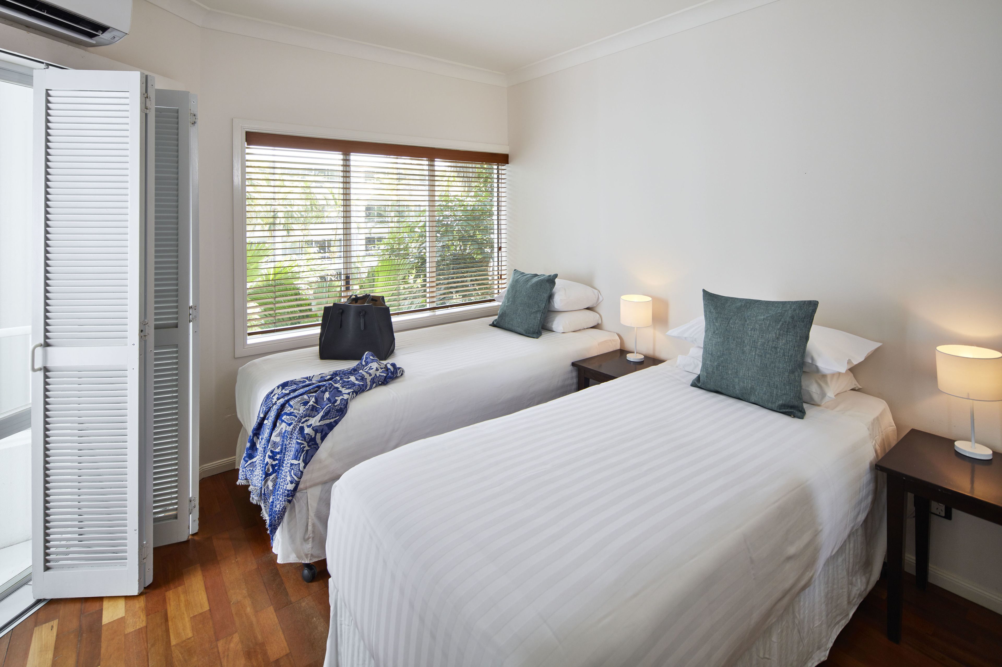 A hotel room with two beds, a window with blinds, a nightstand with a lamp, and a suitcase on one bed.