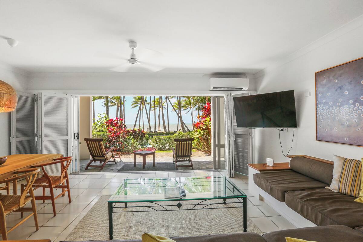 A living room with a glass coffee table, a sofa, and a TV mounted on the wall. There is a door leading to an outdoor area with plants and a view of the beach.