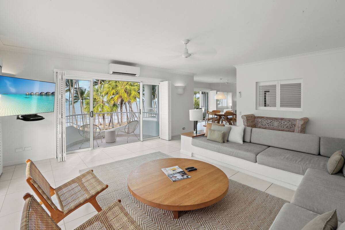 A modern living room with a large TV, a round wooden coffee table, a sofa, and a balcony with a view of palm trees and the sea.