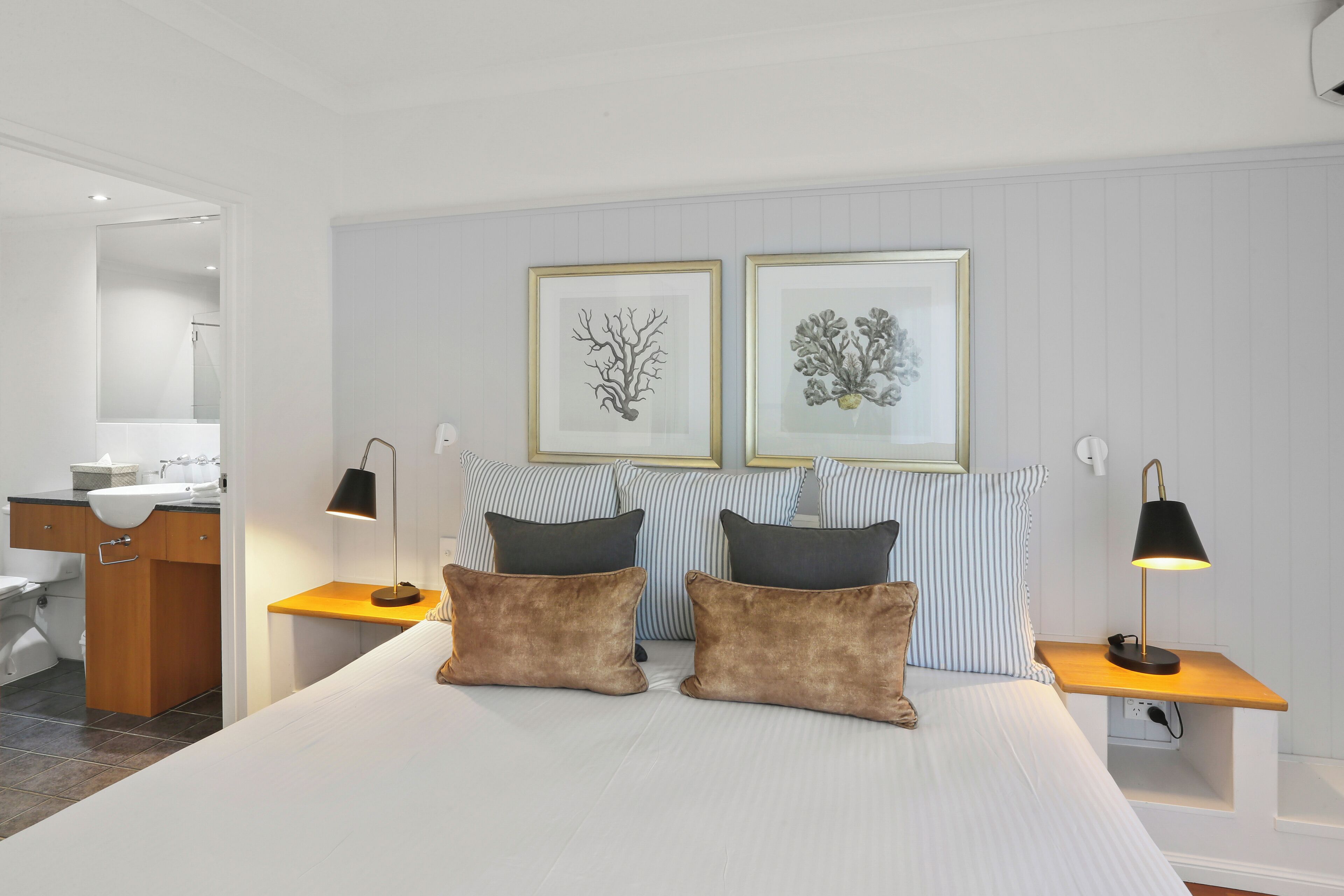 Un hotel moderno con una cama grande, dos mesitas de noche, un tocador, un aparador de madera, un baño y dos cuadros enmarcados en la pared.