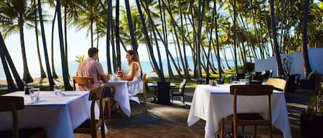 A wooden deck with tables and chairs, palm trees, and a view of the ocean.
