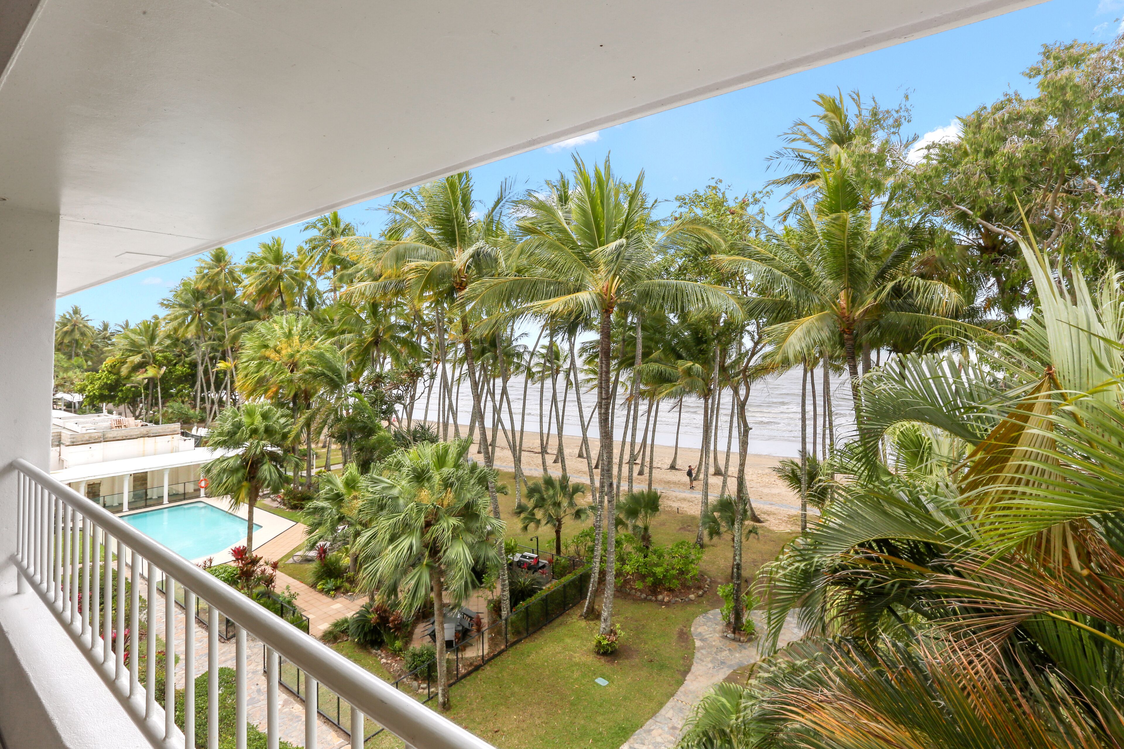Vista de uma varanda com vista para uma área de piscina, palmeiras e uma praia.