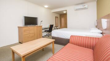 A hotel room with a bed, a desk with a computer, a television, a wooden dresser, and a red-striped sofa.