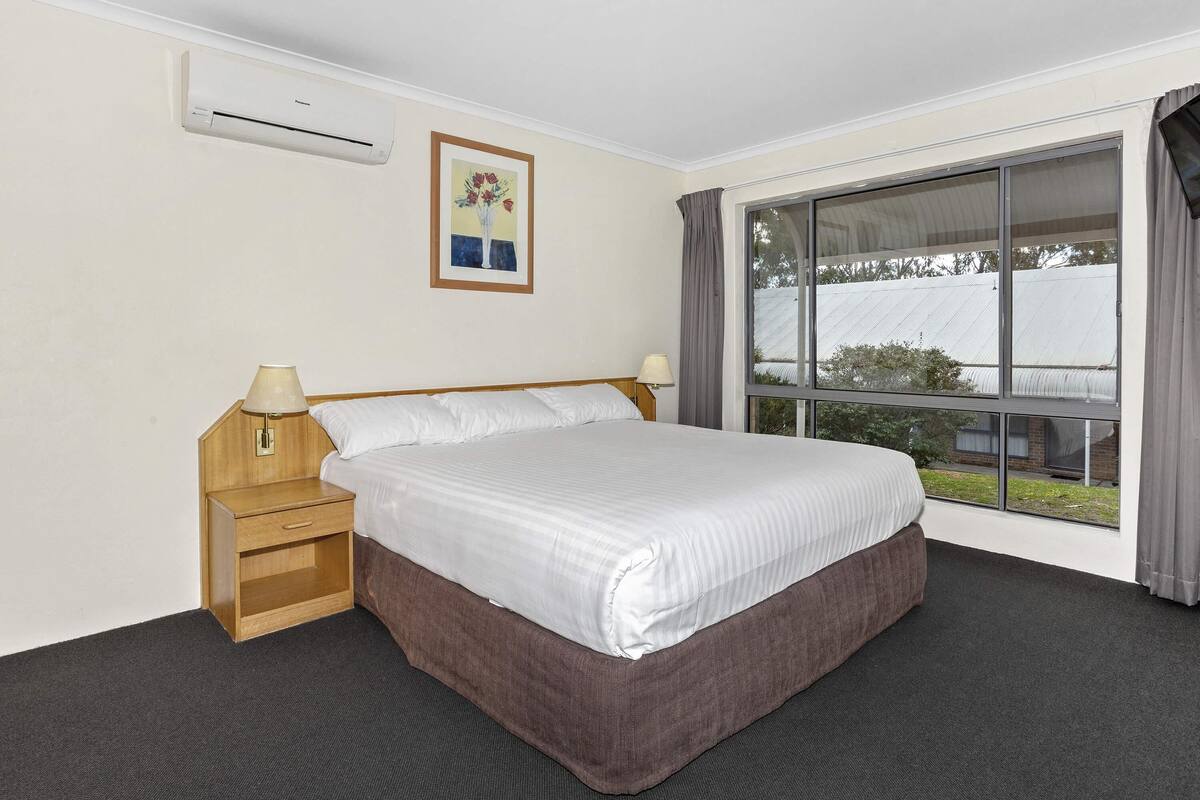 Une chambre d’hôtel avec un lit, une table de chevet, une lampe, un appareil de climatisation, une fenêtre avec des rideaux et un tableau au mur.