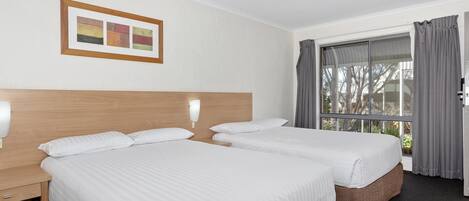 Habitación de hotel con dos camas, cabecera de madera, mesita de noche con lámpara, ventana con cortinas e imagen enmarcada en la pared.
