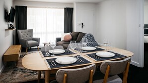 A dining area with a round table set for four, a grey armchair, a sofa, and a TV mounted on the wall. - ibis Styles Canberra Eaglehawk (Sutton)