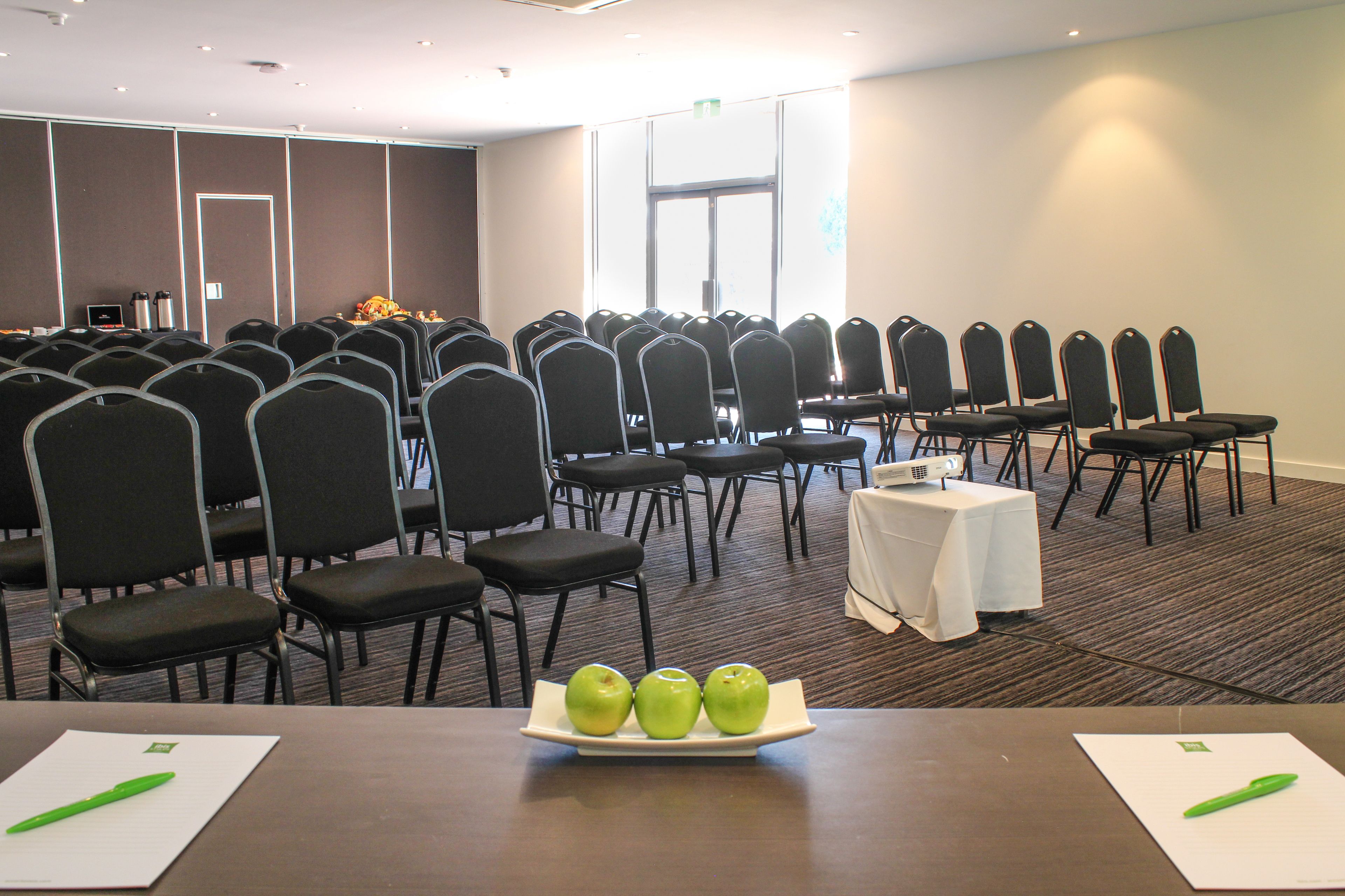 A conference room with rows of chairs, a projector, and a table with apples and pens.