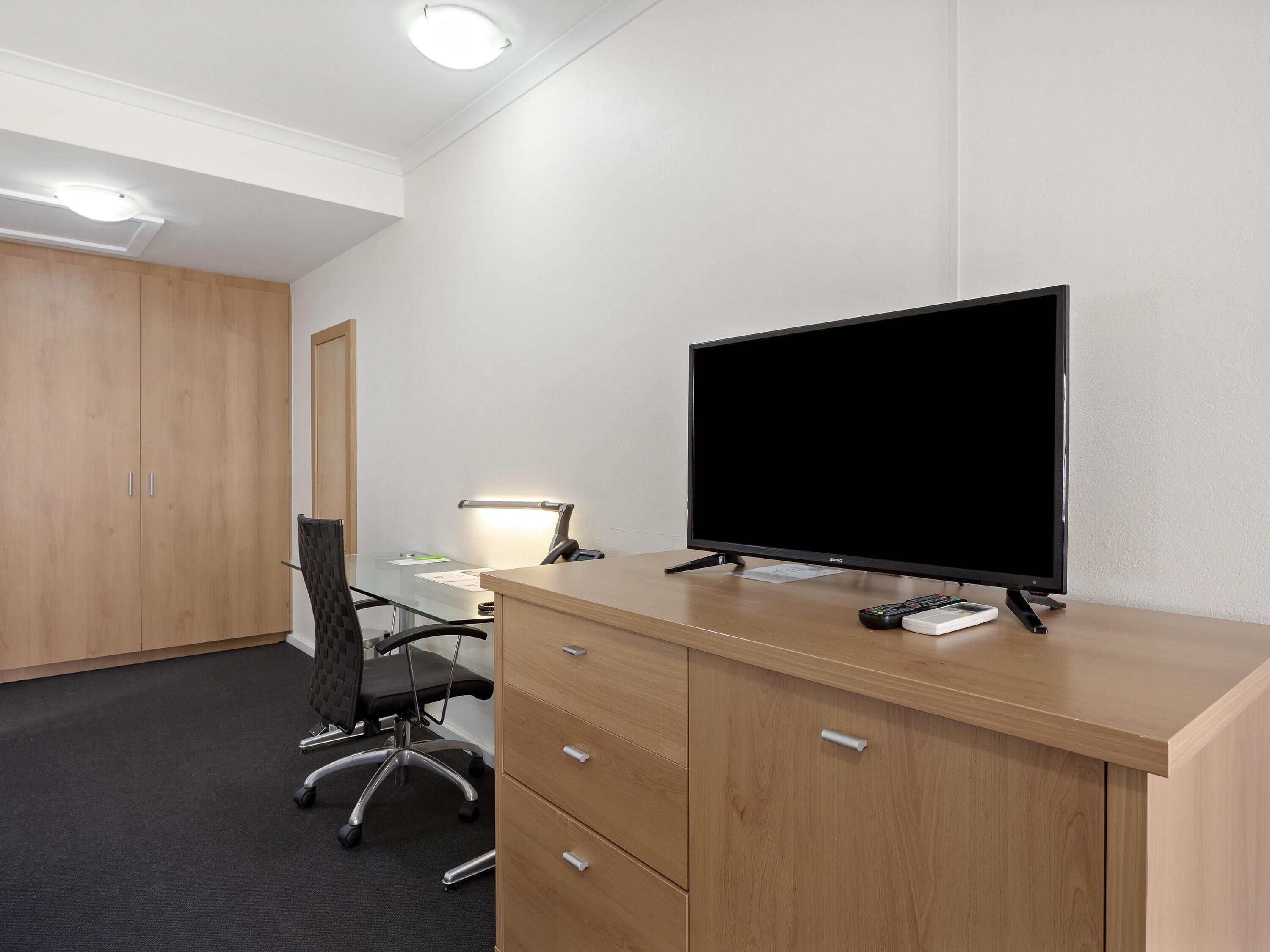 A hotel room with a wooden desk, a black office chair, a flat-screen TV, a desk lamp, and a wardrobe.