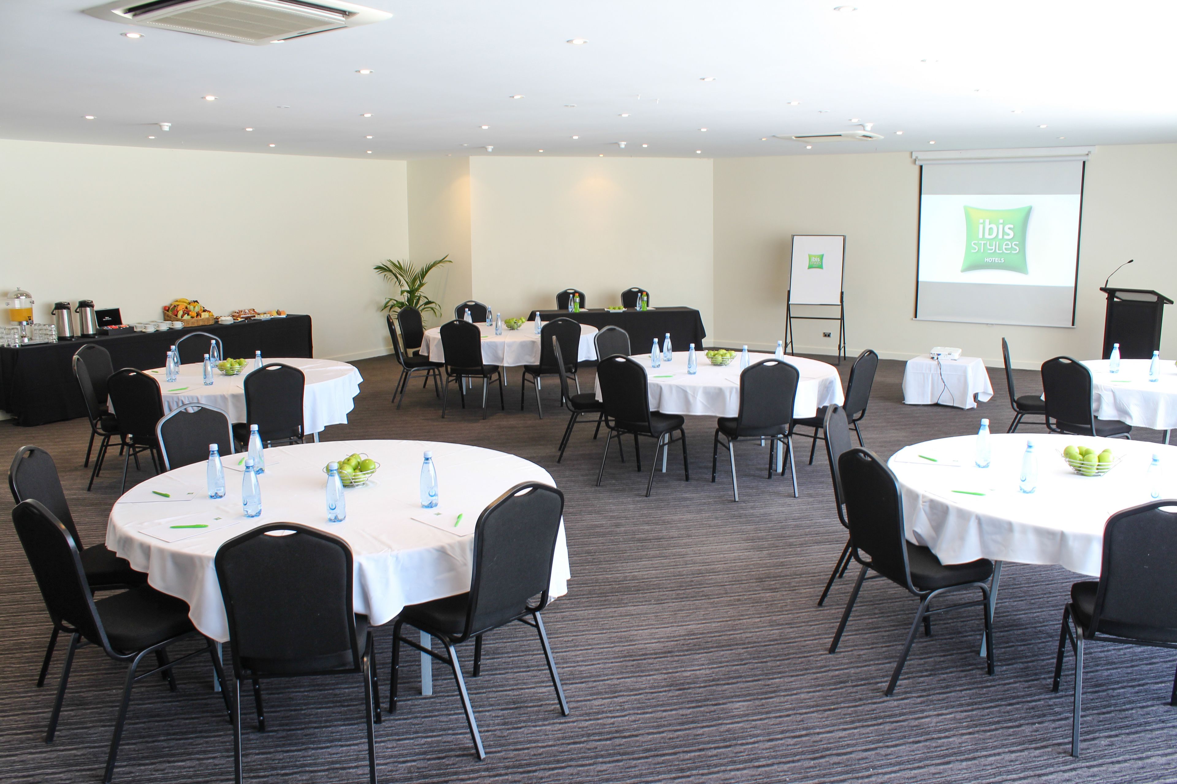 Un salón de conferencias con varias mesas, sillas y una pantalla para presentaciones.