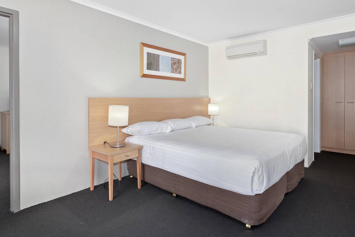 Une chambre d’hôtel avec un lit, des lampes de chevet, une petite table, un tableau encadré au mur et un appareil de climatisation.