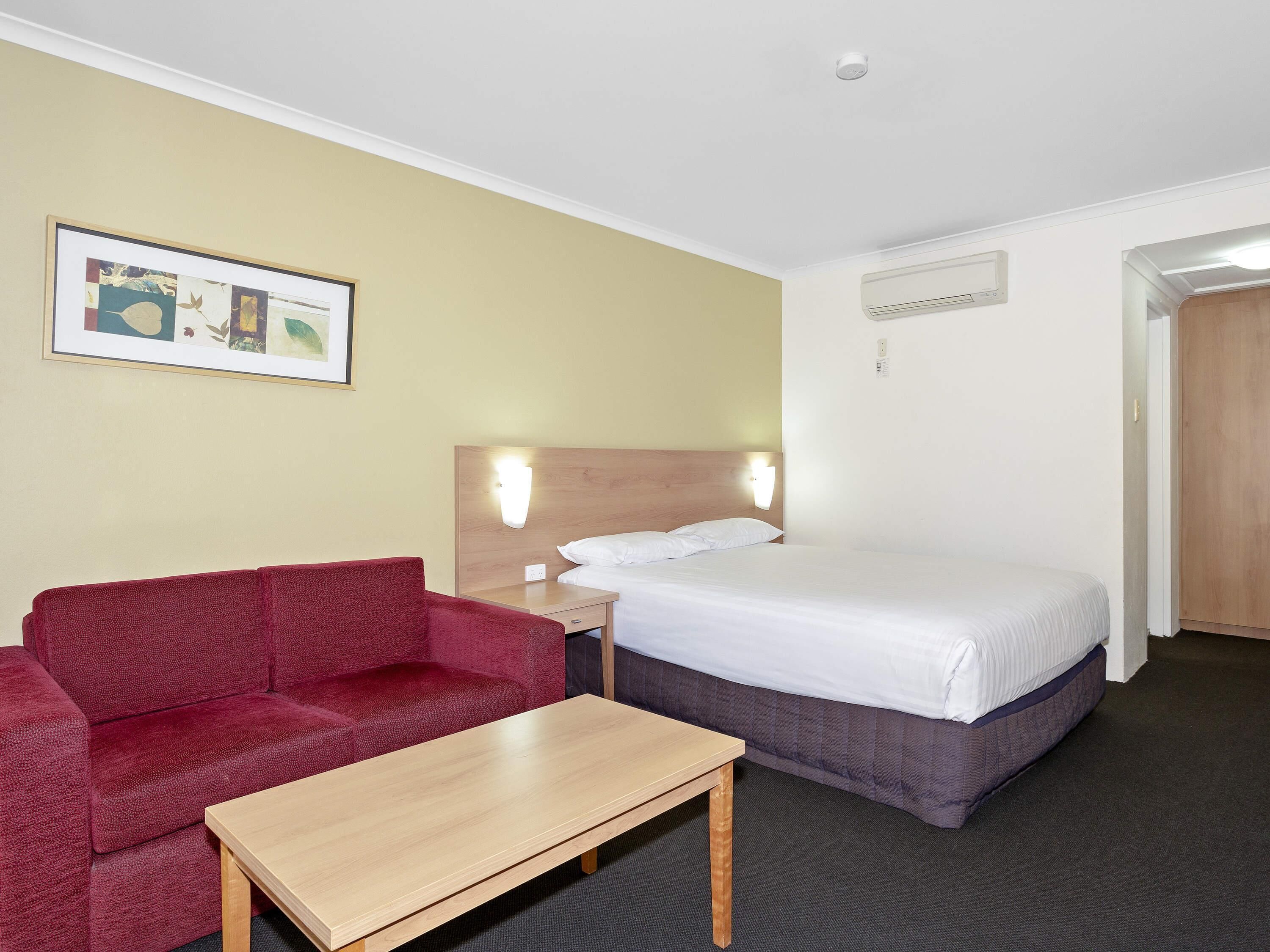Hotellihuone, jossa on sänky, punainen sohva, puinen pöytä, seinään kiinnitetty valaisin ja kehystetty taulu.
