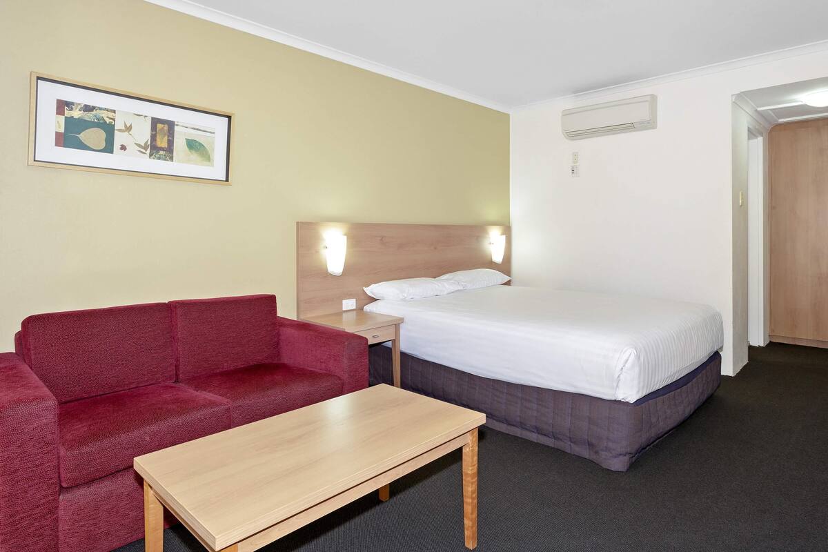 Une chambre d’hôtel avec un lit, un canapé rouge, une table en bois, un lampadaire fixé au mur et un tableau encadré.