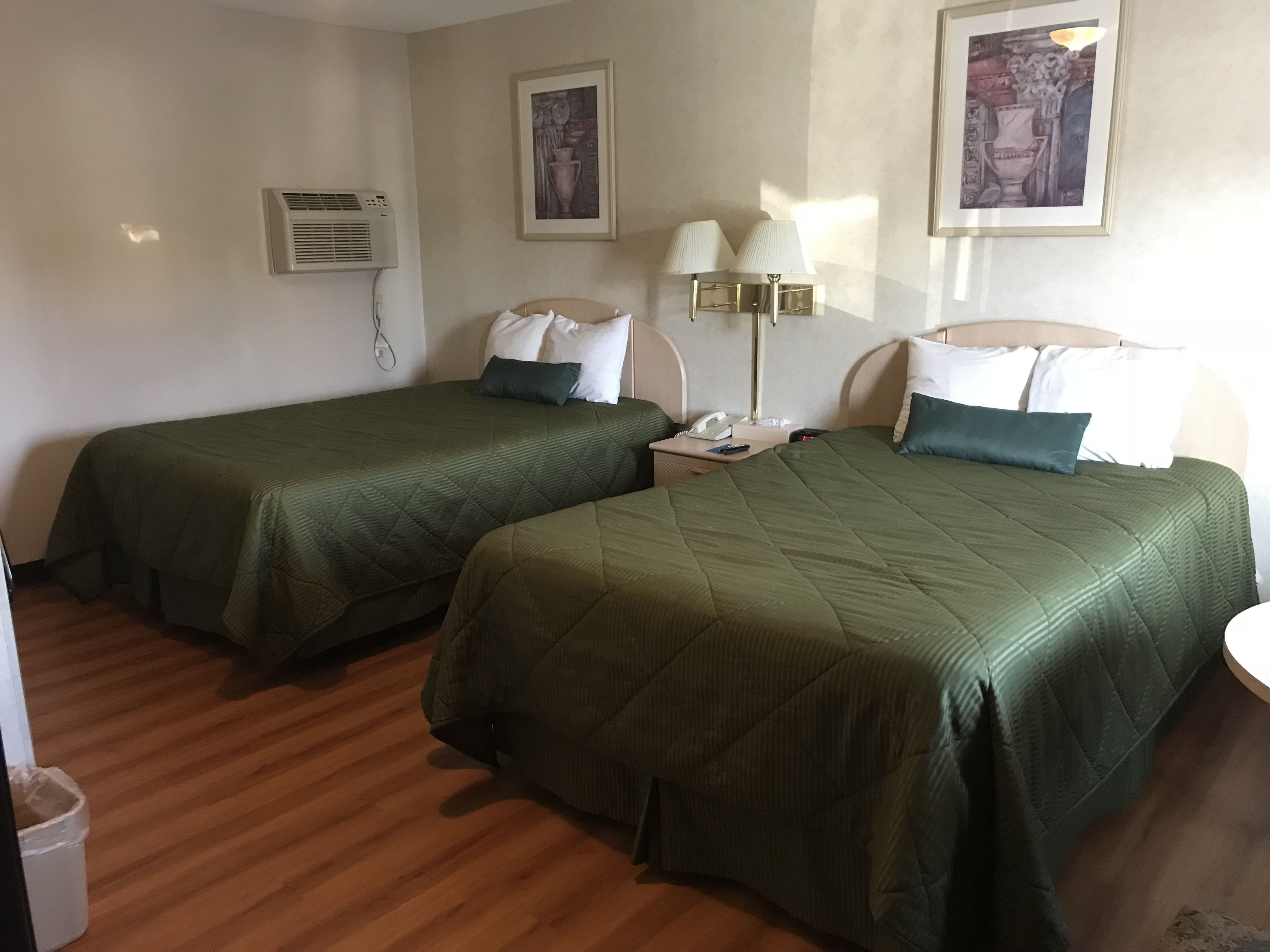 A hotel room with two single beds, a bedside table with a lamp, a wall-mounted air conditioner, and framed artwork on the walls.