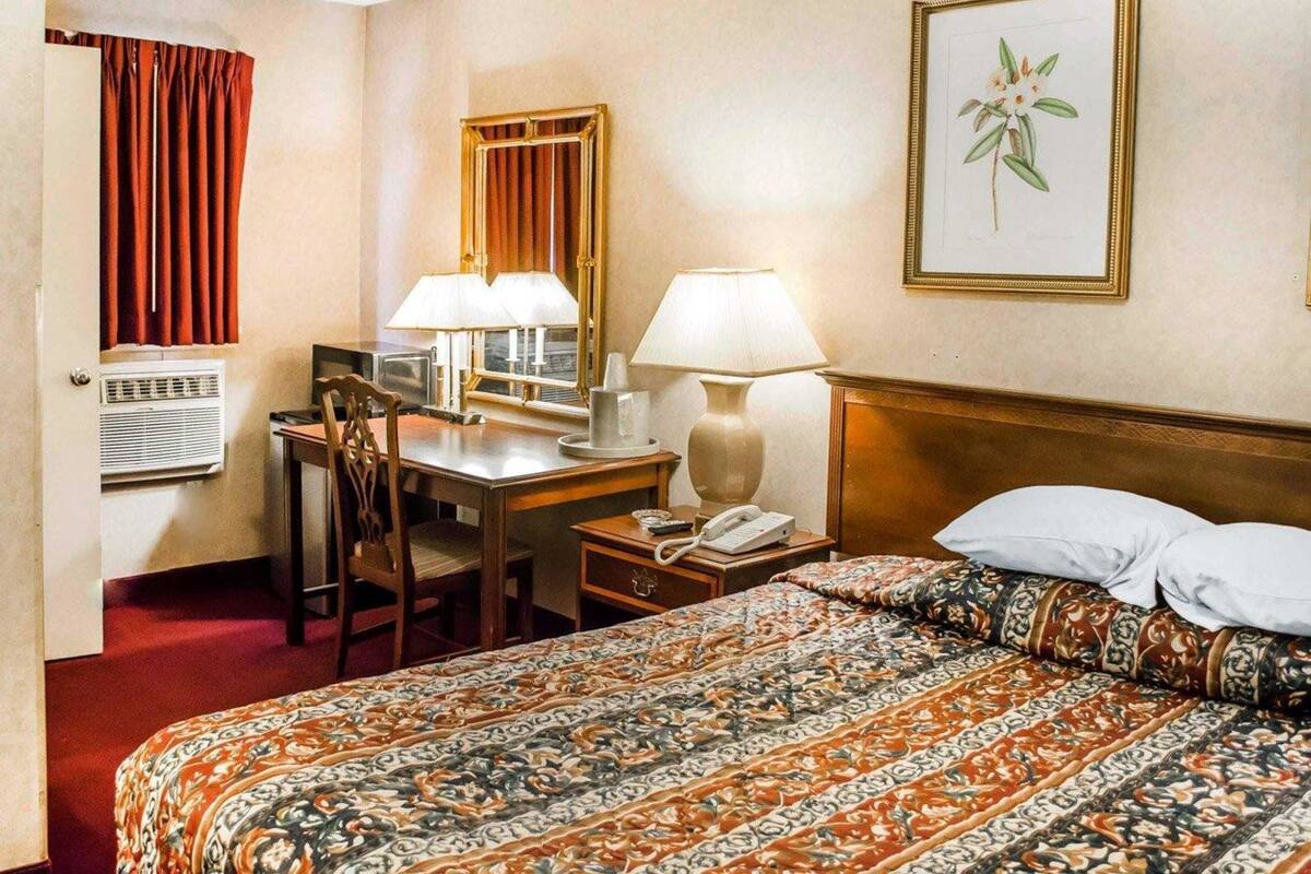 Una habitación de hotel con una cama, un escritorio con dos lámparas, un teléfono, una silla y un ventanal con cortinas.