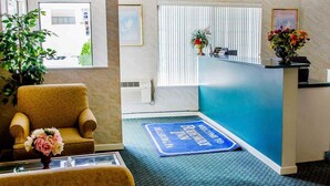 A hotel reception area with a reception desk, a blue welcome mat, a chair with a vase of flowers, and a potted plant.
