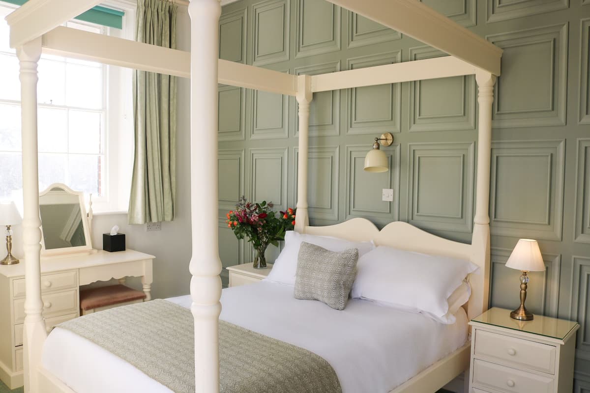 Ein Bett mit Himmel, ein weißes Kopfteil, ein Nachttisch mit Lampe und ein Beistelltisch mit Blumen.