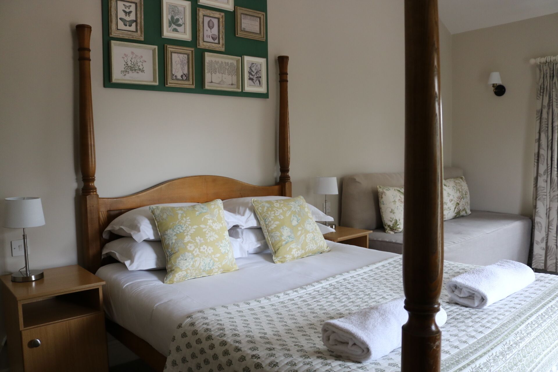 Un lit à baldaquin agrémenté de coussins à motifs floraux, une table de chevet avec une lampe et un mur orné d’illustrations botaniques encadrées.