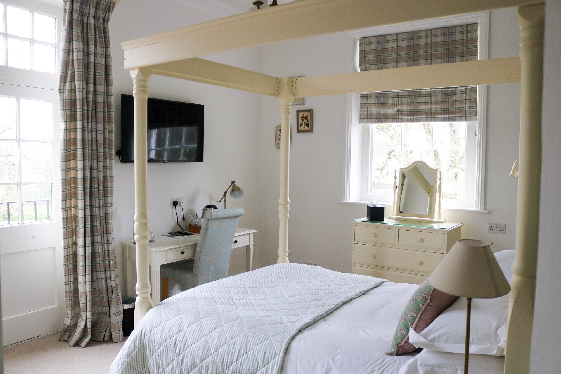 A four-poster bed with a quilted duvet, a bedside lamp, a dresser with a mirror, and a desk with a lamp.