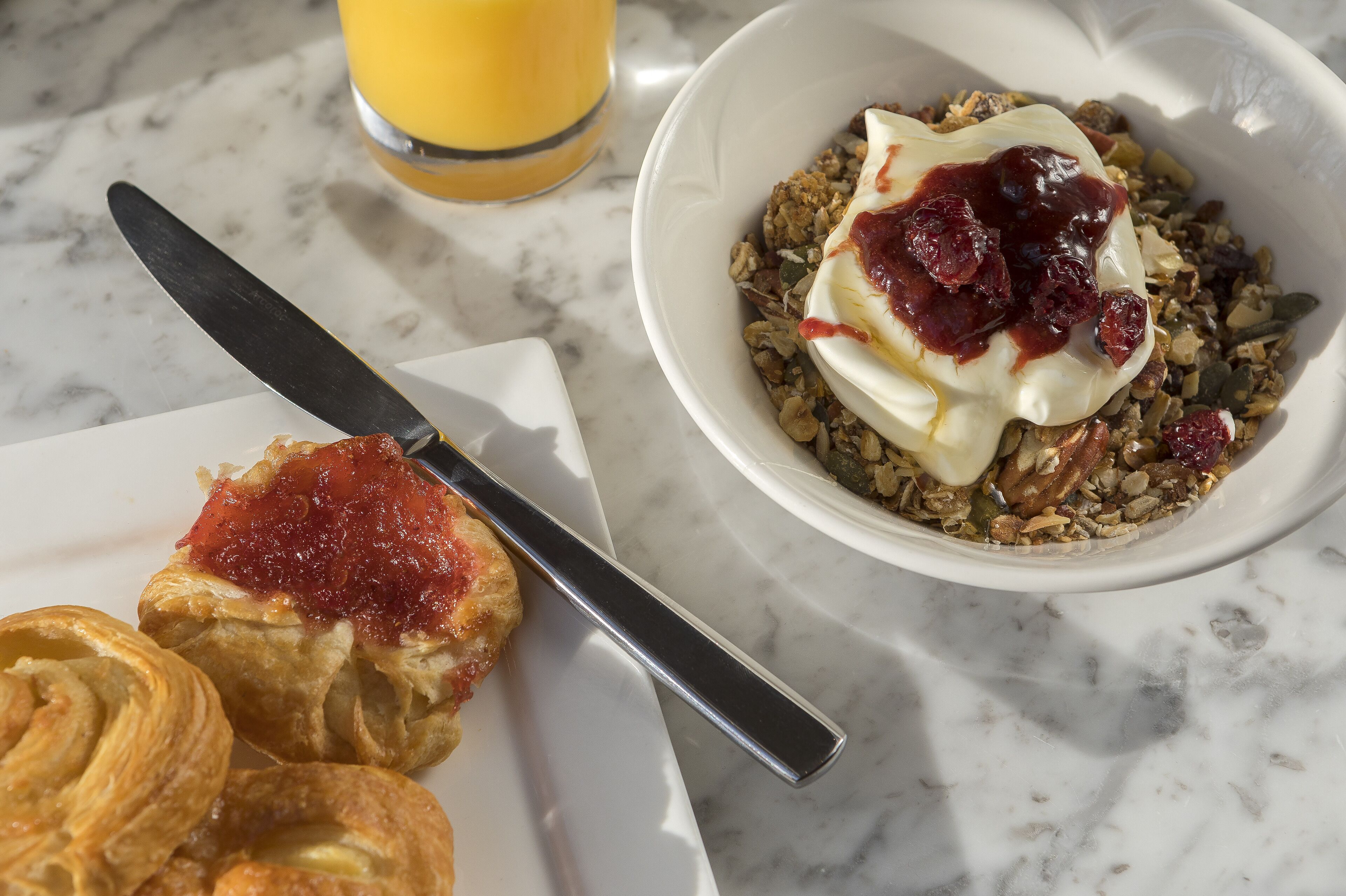 A breakfast setting with a bowl of cereal topped with yogurt and berries, a plate with pastries, and a glass of orange juice on a marble table.