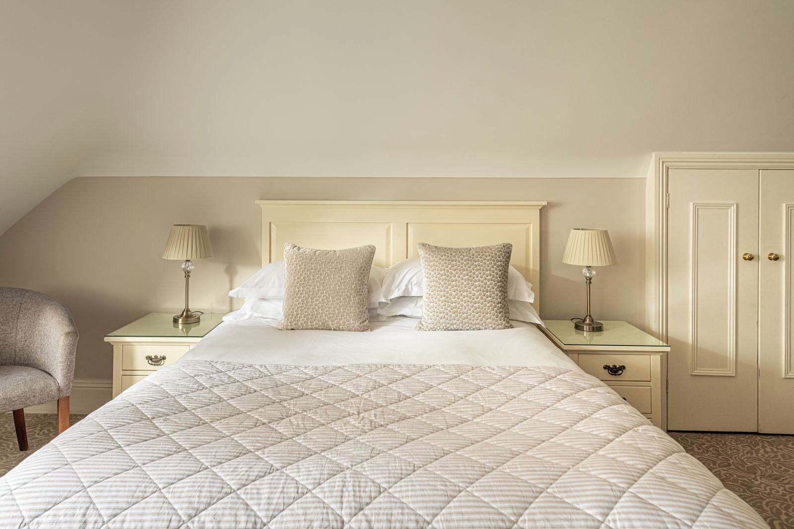 A neatly arranged bedroom with a large bed, two bedside tables with lamps, a chair, and a wardrobe.