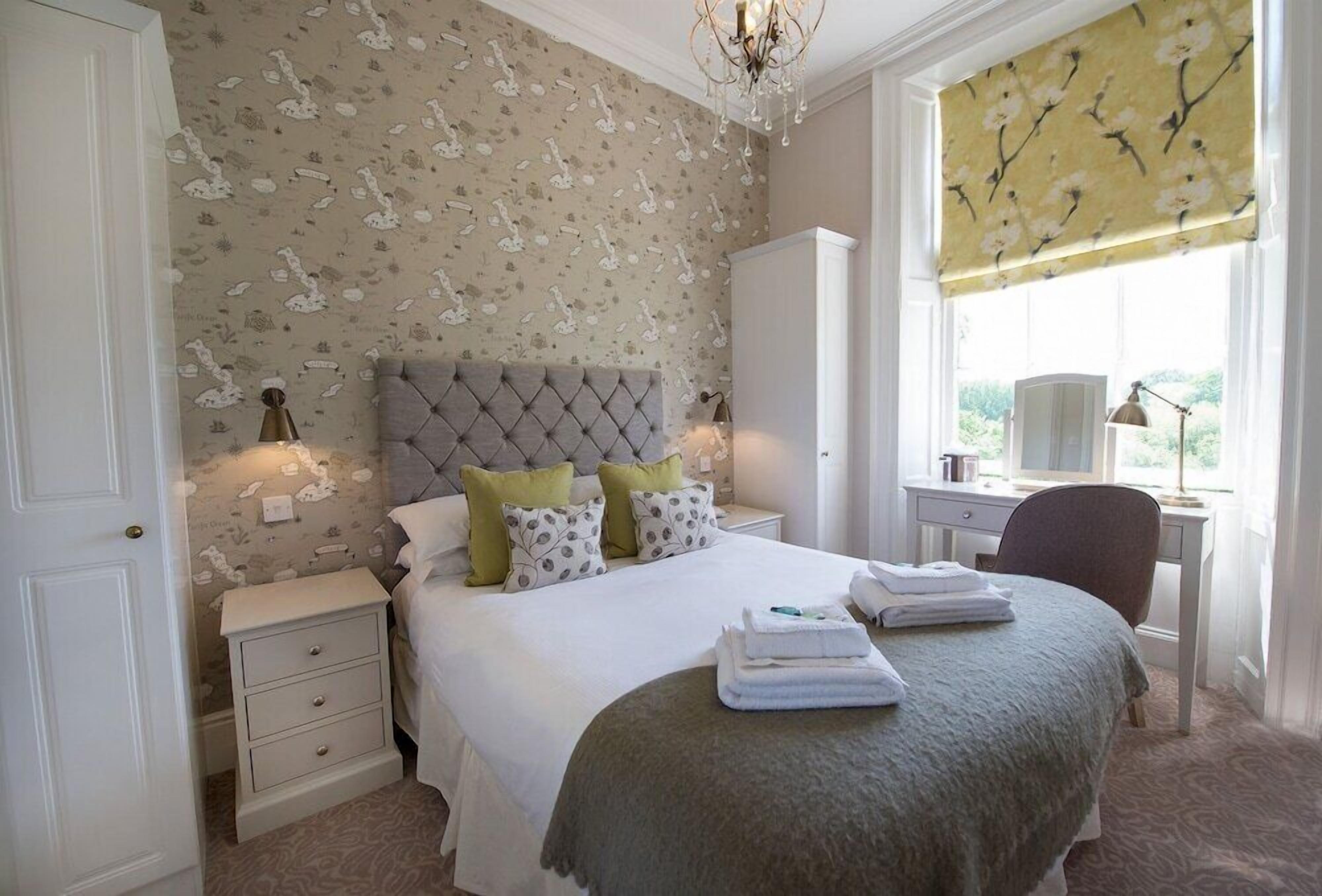 A bedroom with a tufted headboard, a bed with white linens and green pillows, a nightstand with drawers, a desk with a lamp, and a window with a floral blind.