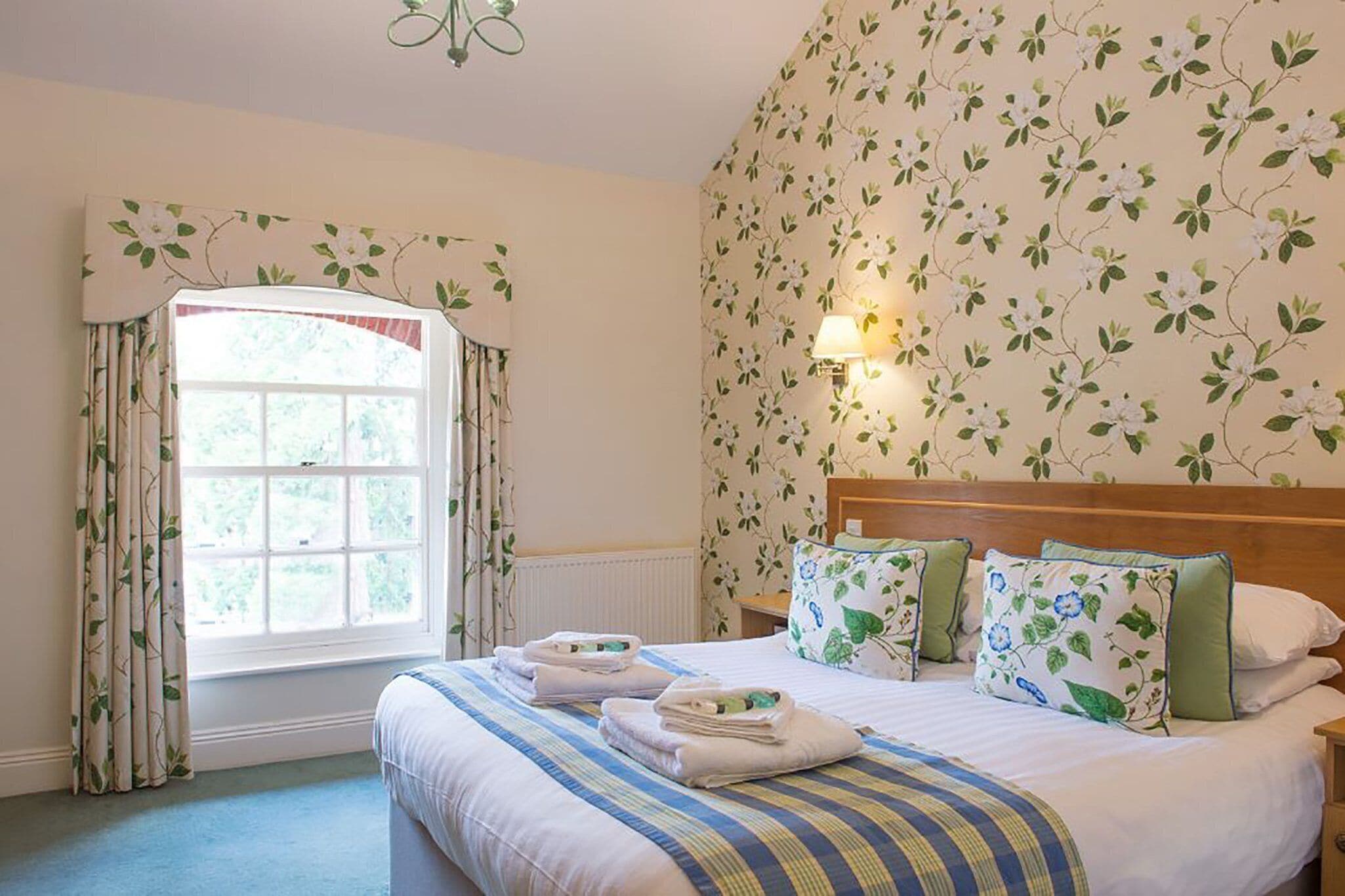 A bedroom with a large window, a bed with striped bedding, floral-patterned pillows, and a bedside lamp.