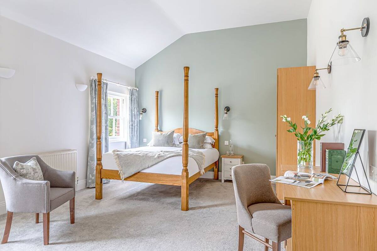 Ein Schlafzimmer mit Himmelbett, Schreibtisch mit Stuhl, Nachttisch, Kommode und einem Fenster mit Vorhängen.