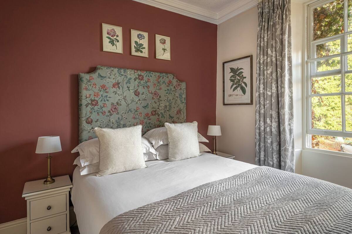 Ein ordentlich bezogenes Bett mit weißen Leinen und einer gemusterten Tagesdecke, ein Blumen-Kopfstück und gerahmte Pflanzenillustrationen an der Wand.