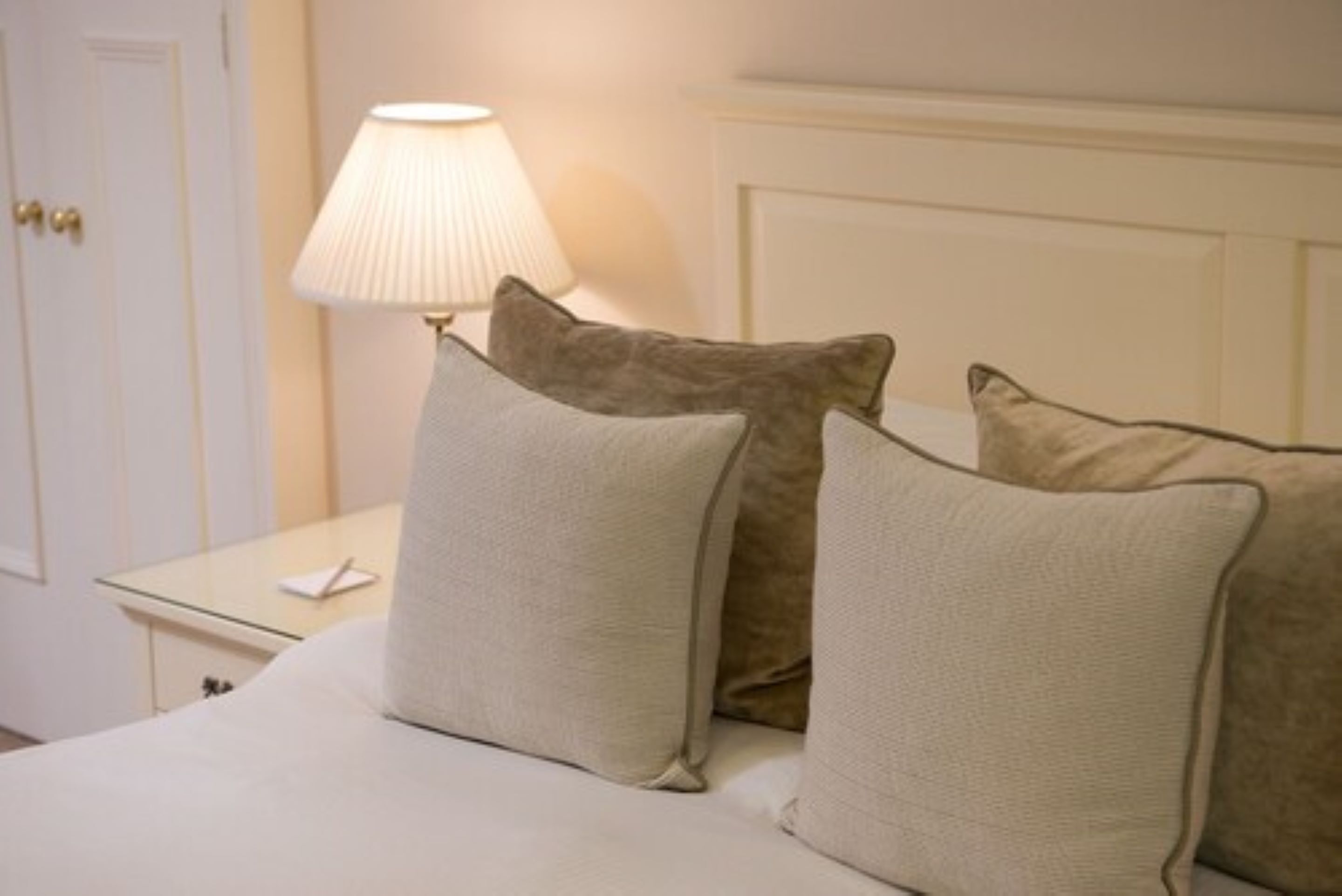Una cama bien hecha con almohadas color beige y una lámpara blanca sobre una mesita de noche.