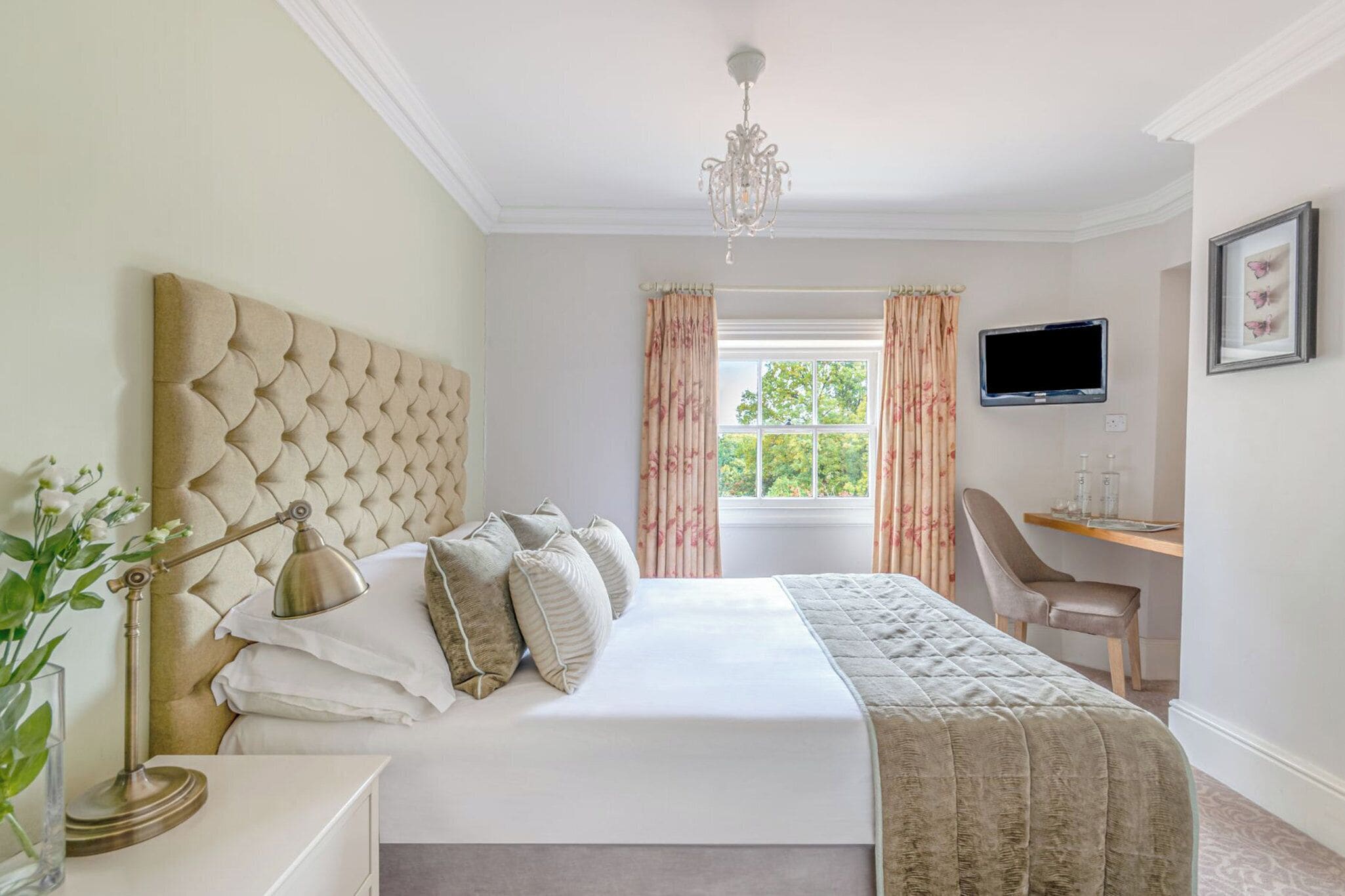 A bedroom with a tufted headboard, a bedside lamp, a bed with white linens, a desk with a chair, a television, and a window with curtains.