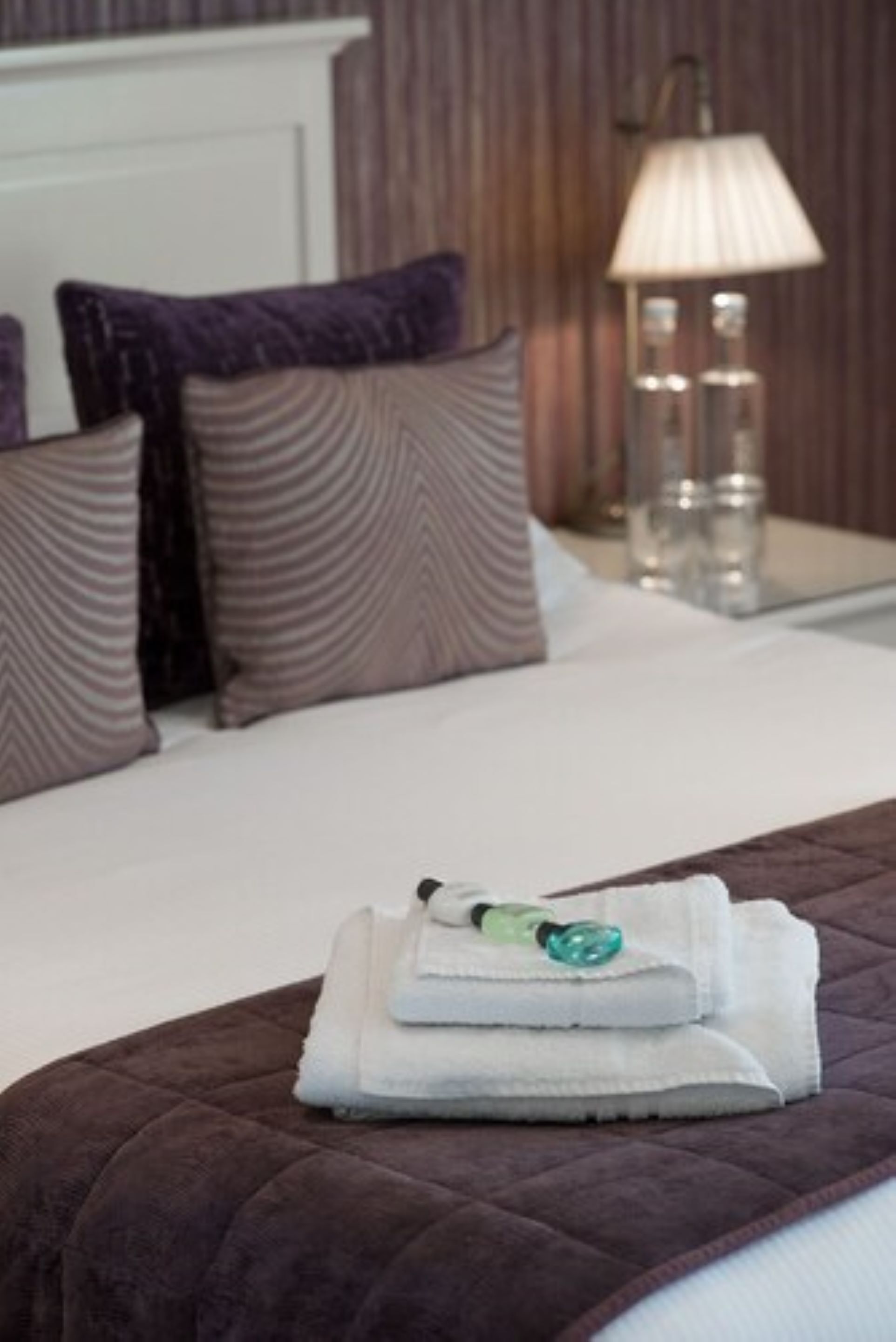Un lit bien fait, recouvert d’une couverture marron, avec une serviette blanche pliée et une bouteille verte posée dessus.