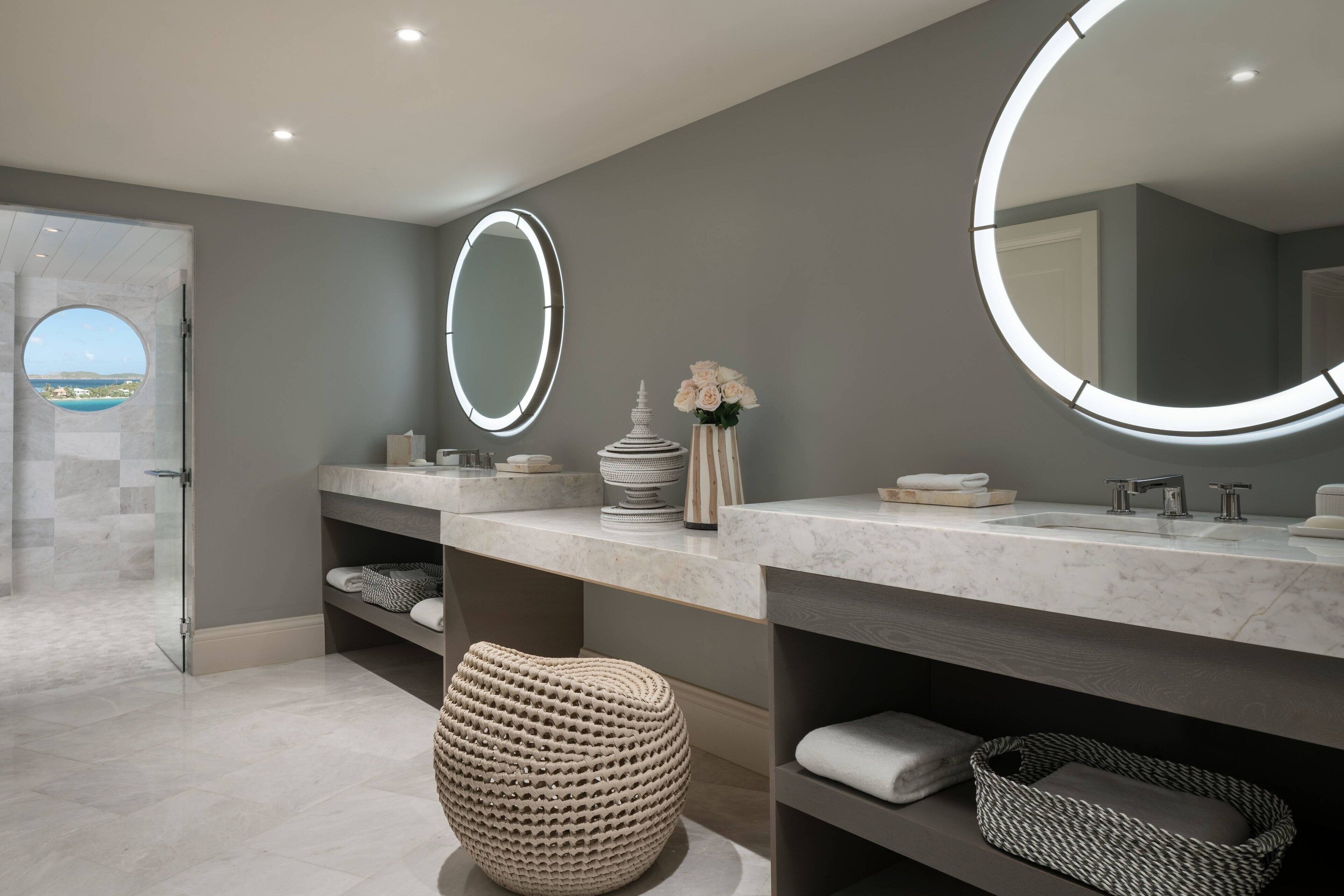 Une salle de bain moderne avec deux lavabos, de grands miroirs et une vue à travers une porte en verre.