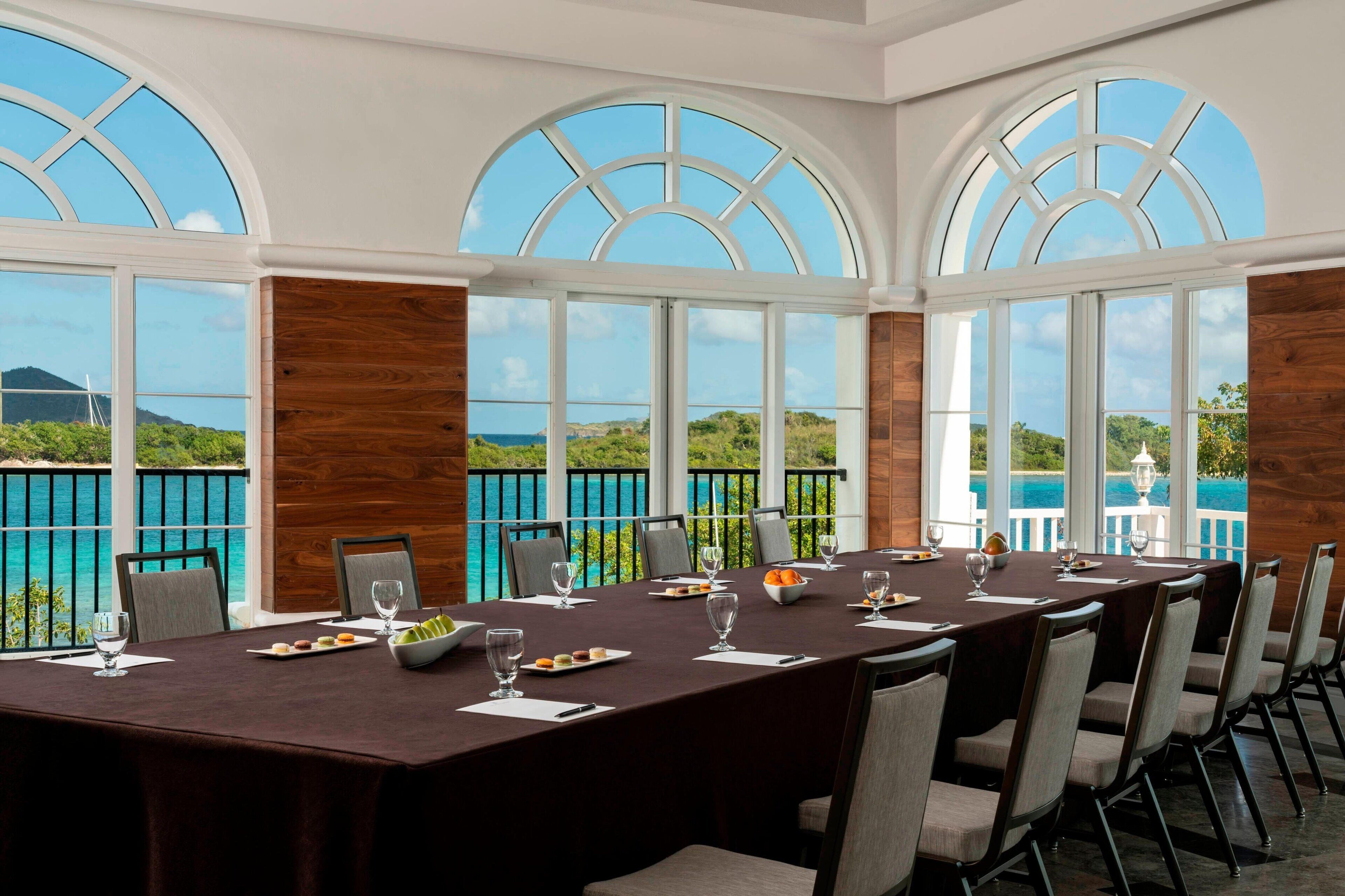 A conference room with a long table set for a meeting, large arched windows, and a view of the ocean.