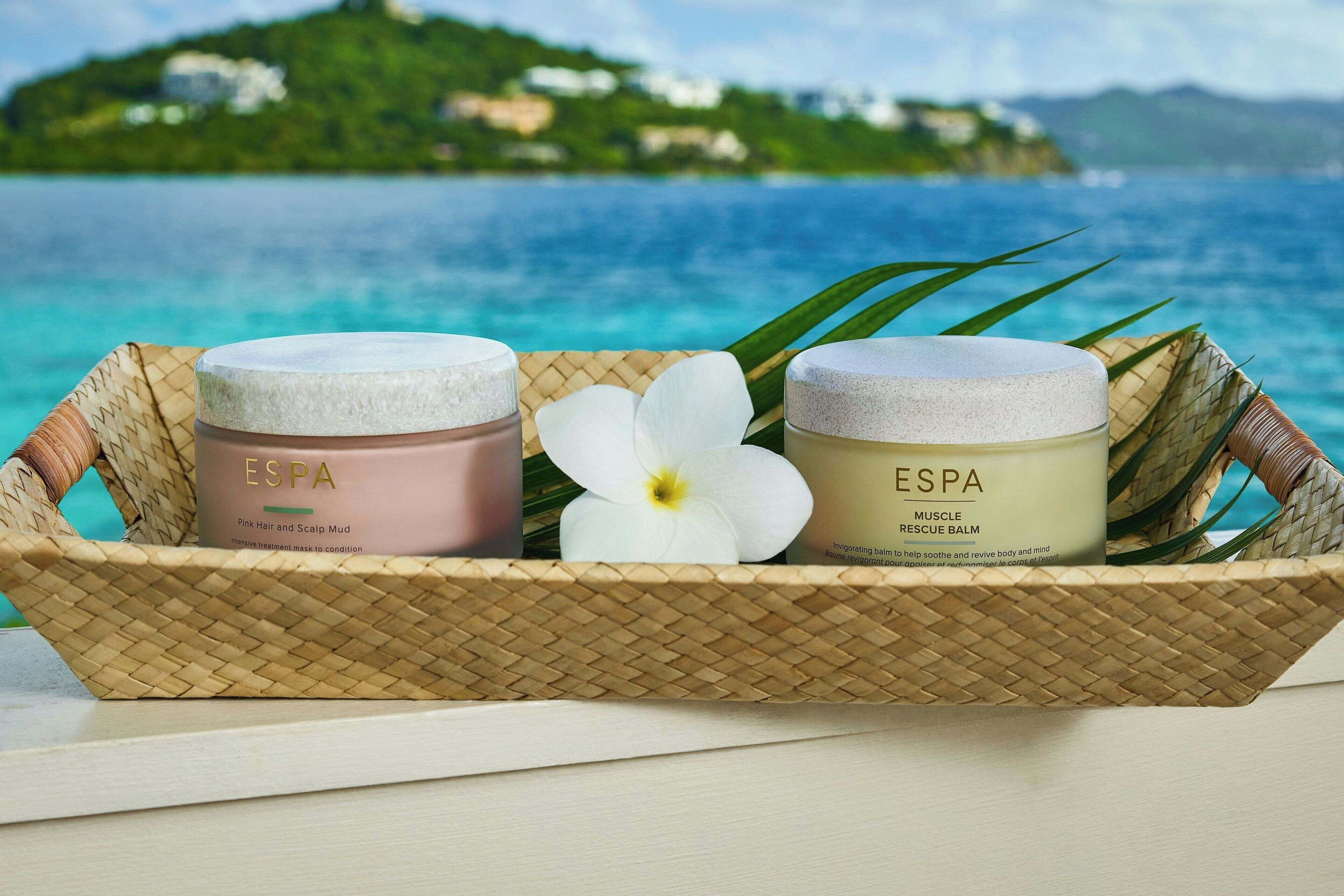 A wicker basket with two jars of ESPA products and a white flower on a table with a view of a beach and ocean.