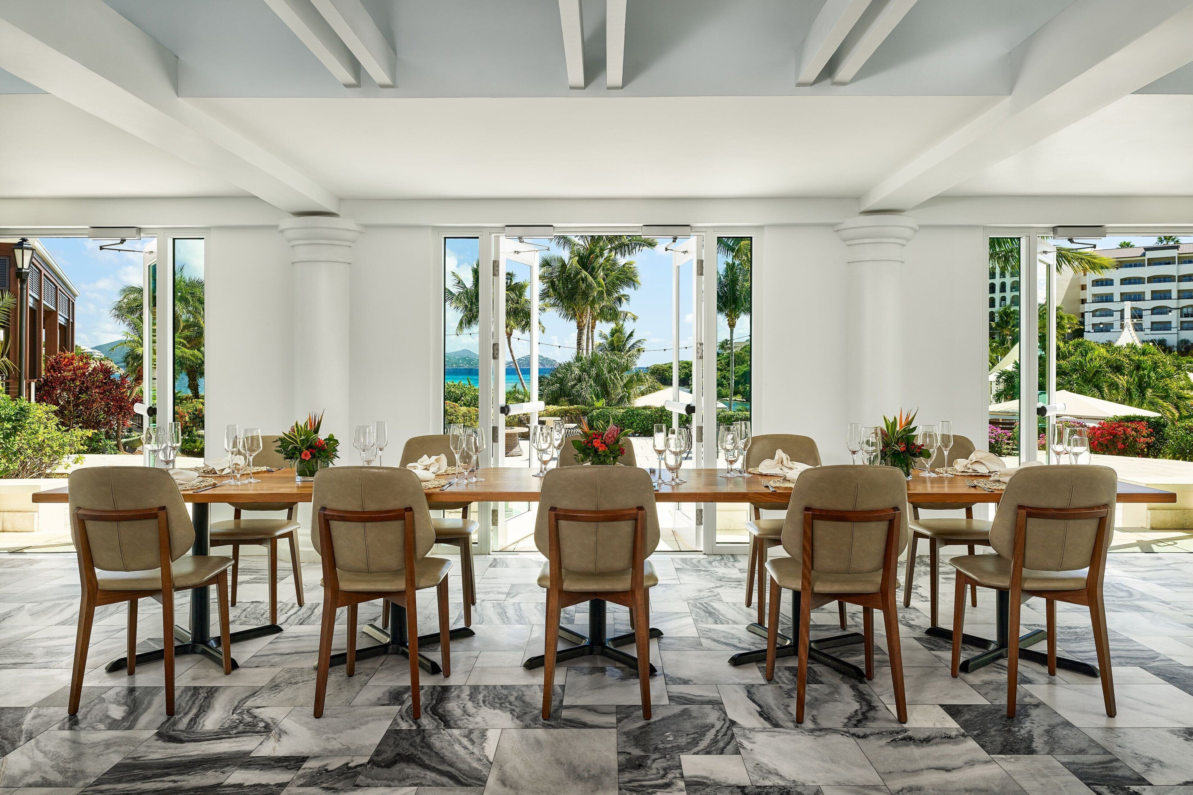 A dining area with a large table set for six, surrounded by six chairs, with a view of palm trees and a building in the background.