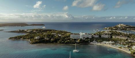 An aerial view of a coastal resort with multiple buildings, a marina with yachts, and clear blue waters.