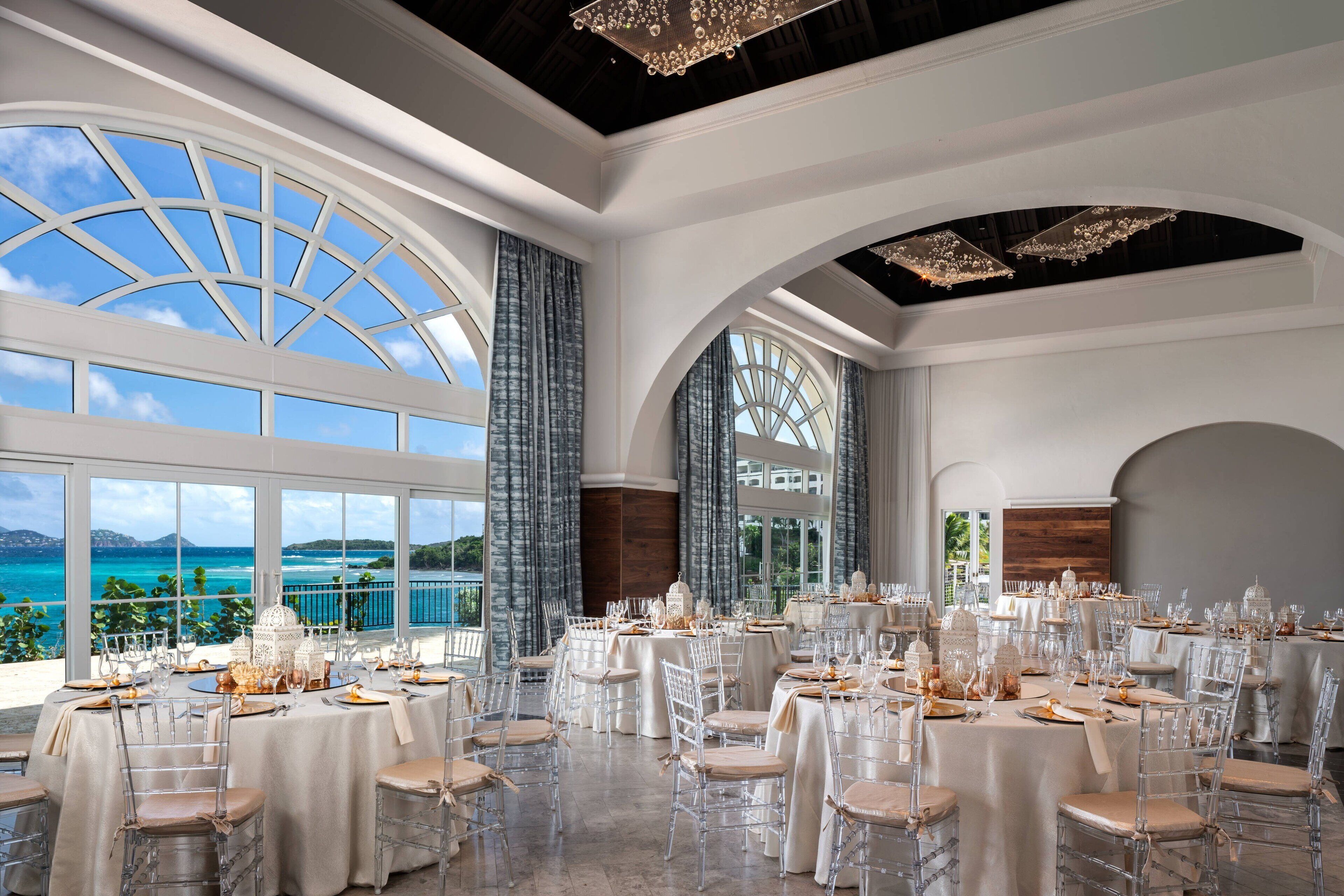 Una sala banchetti con tavoli rotondi, sedie trasparenti e ampie finestre vista mare.