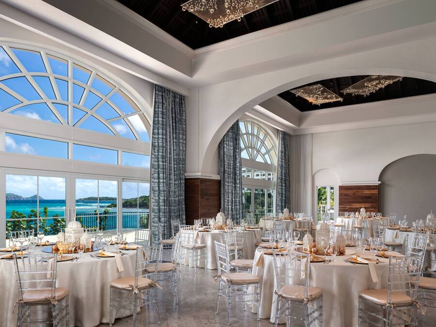 A banquet hall with round tables, clear chairs, and large windows offering a view of the sea.