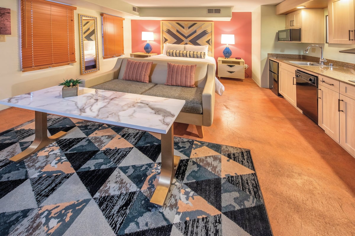 A compact hotel room with a bed, a dining table, a sofa, a kitchen area, and a patterned rug.