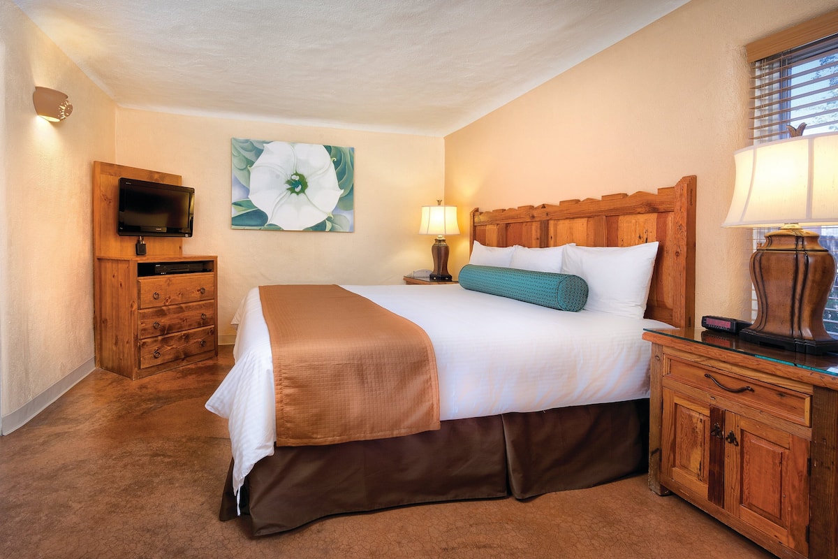 A hotel room with a bed, a television, a wooden nightstand, a lamp, and a painting on the wall.
