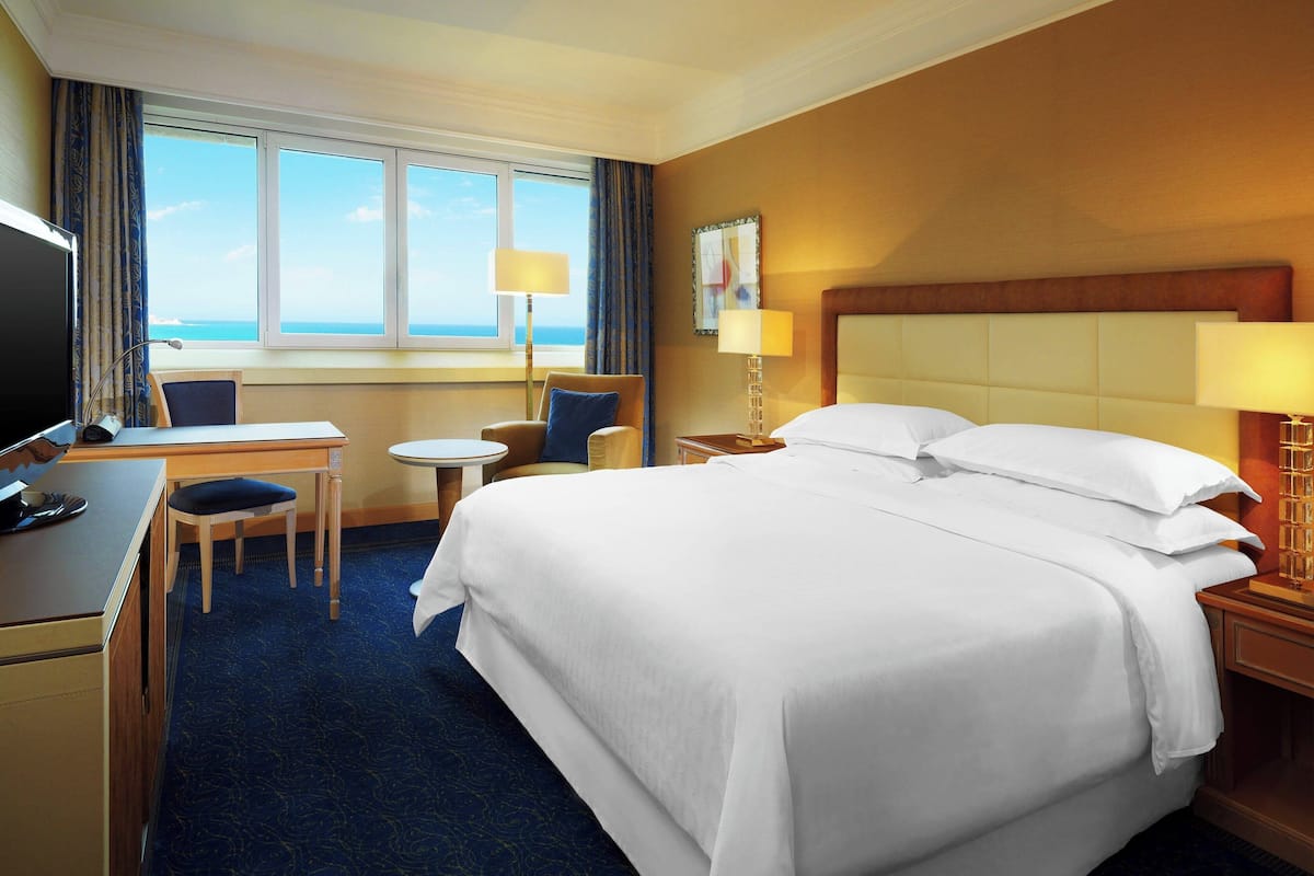 Ein Hotelzimmer mit einem großen Bett, einem Schreibtisch, einem Sessel, einem Fernseher und Blick auf das Meer.