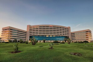 A modern hotel with a curved facade, multiple floors, and a well-maintained lawn with small plants.