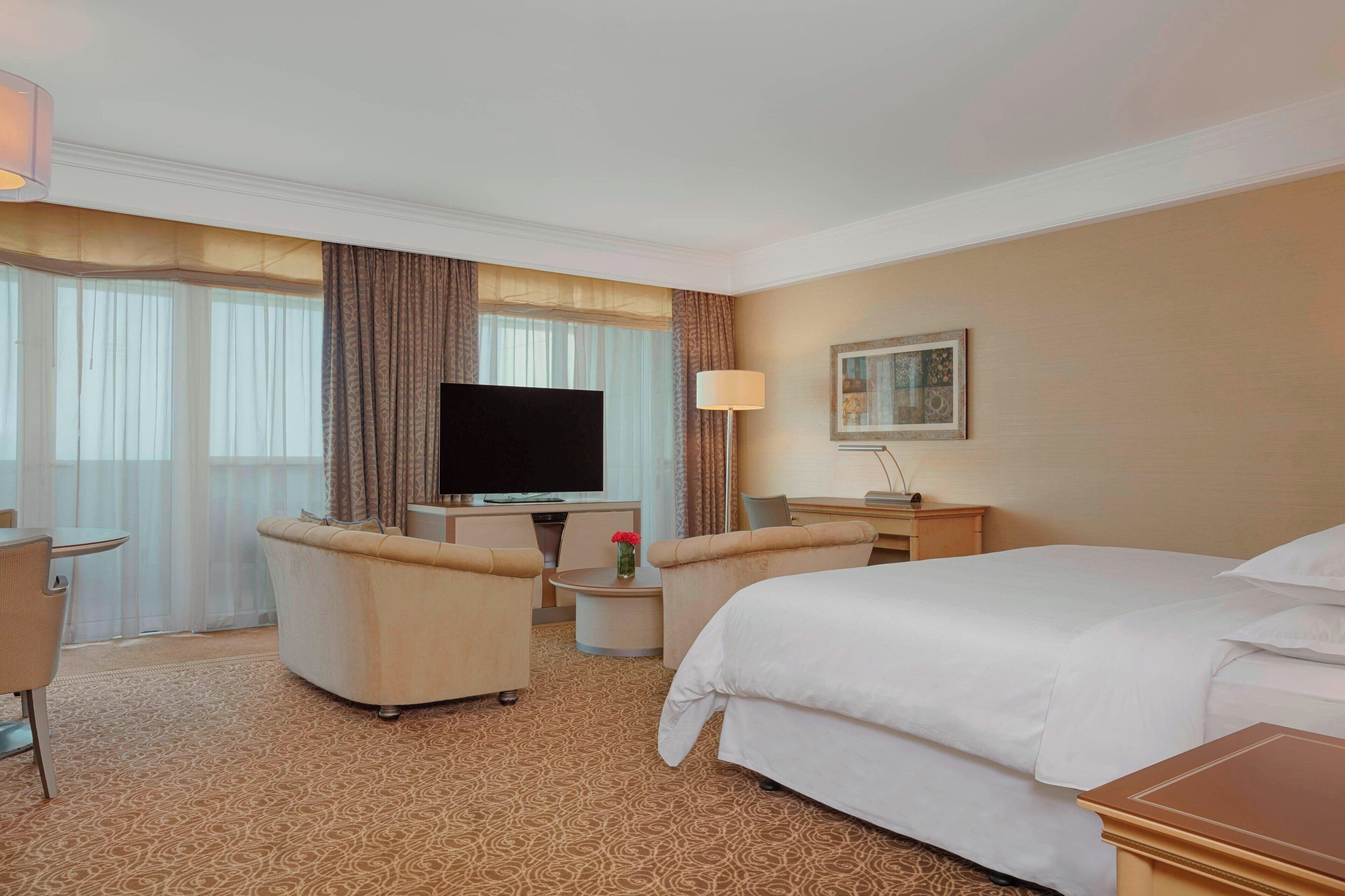 A hotel room with a large bed, a flat-screen TV, a desk with a chair, and a small table with a vase of flowers.