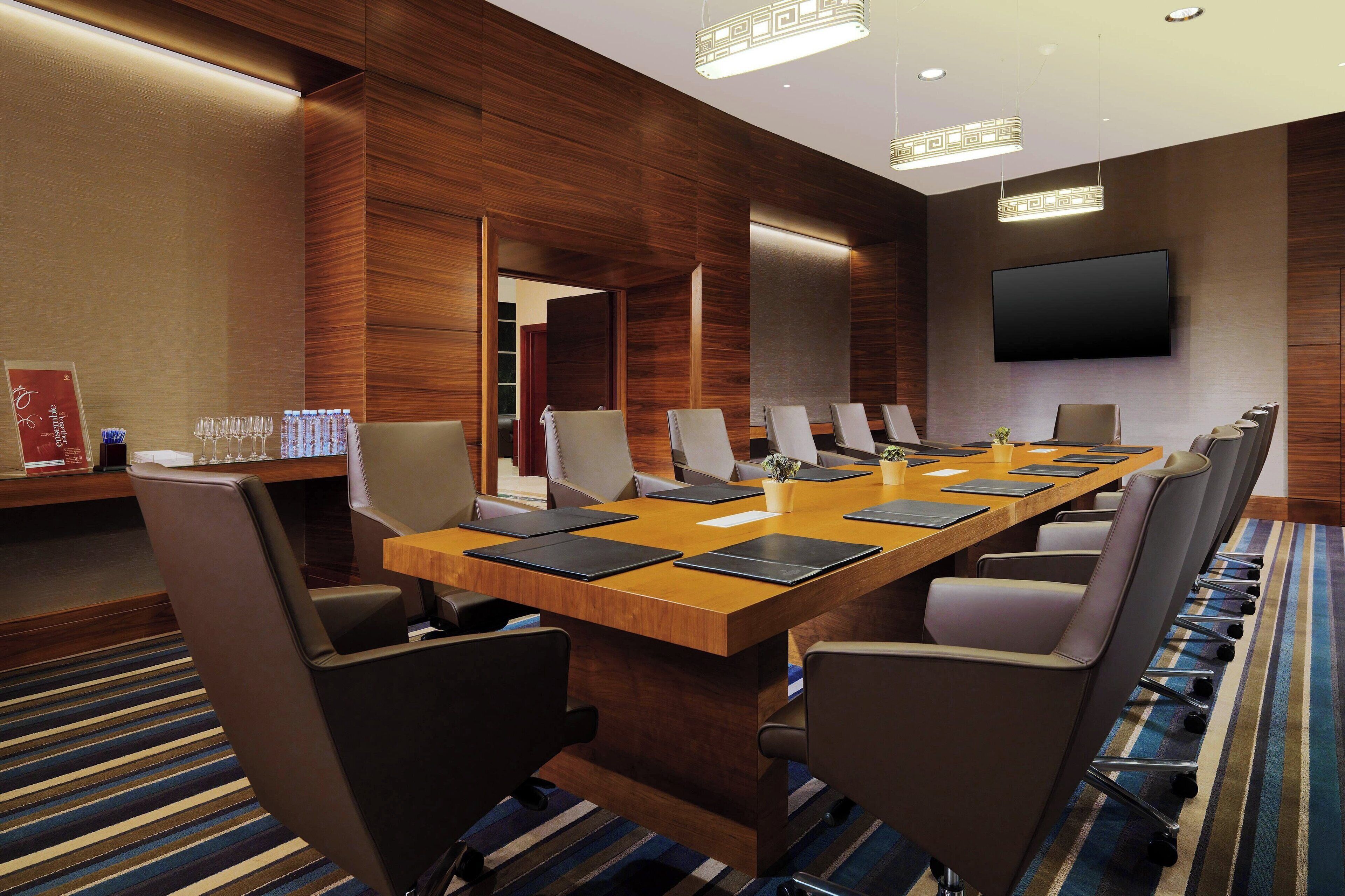 A conference room with a long table, multiple chairs, a flat-screen TV, and wooden paneling.