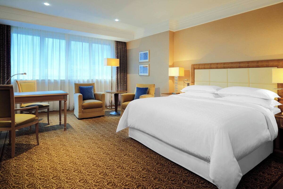 Ein Hotelzimmer mit einem großen Bett, einem Schreibtisch, zwei Stühlen, einem kleinen Tisch und zwei Lampen.