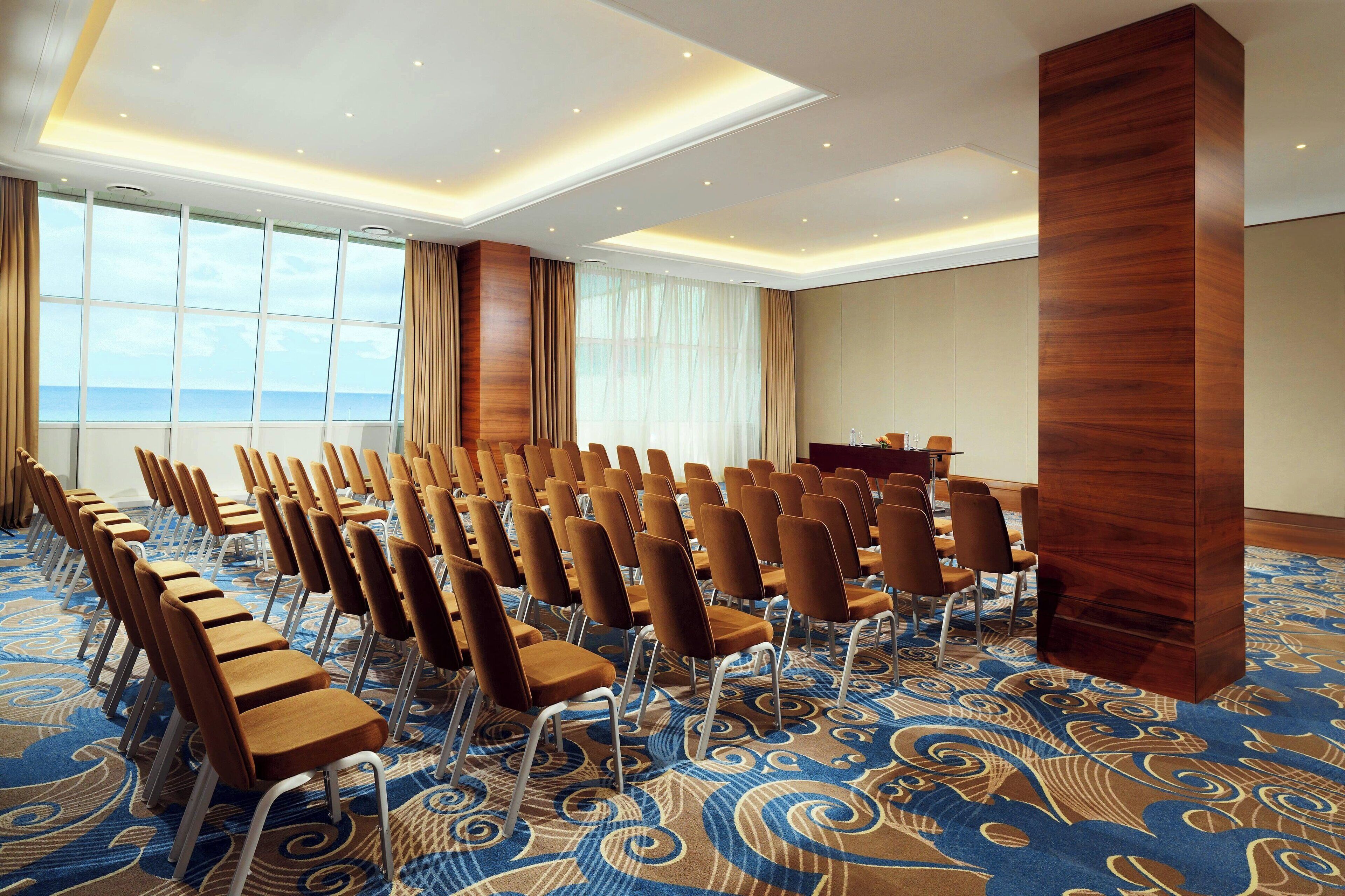 Une salle de conférence avec des rangées de chaises, une grande colonne en bois et un tapis à motifs.