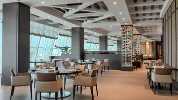 Een modern restaurant met ronde tafels en stoelen, grote ramen en een centrale kolom met een wijnrek.