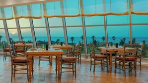 A restaurant with large windows offering ocean views, wooden tables and chairs, and a tiled floor. - Sheraton Club des Pins Resort (Algiers)